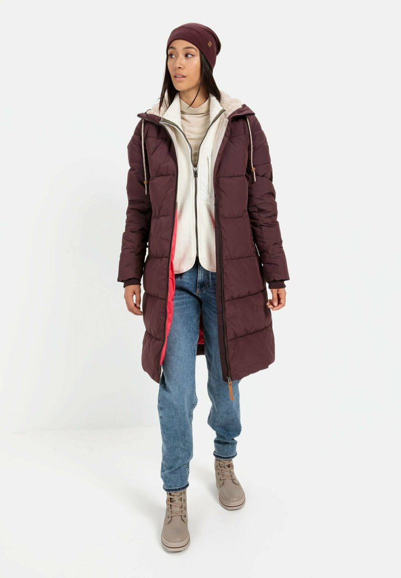 Bordeaux puffer jas met capuchon en witte fleece voering, gelaagd over een lichtbeige top, gecombineerd met blauwe jeans en beige laarzen.