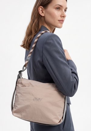 Frau trägt ein graues Sakko und eine beige GMV-Schultertasche mit einem gemusterten Stoffriemen über der Schulter.