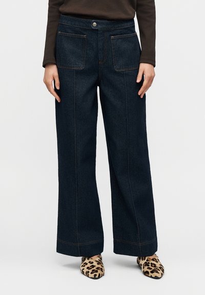 Jean en denim foncé à jambes larges avec deux poches avant, couture orange contrastante et taille haute. Chaussures à imprimé léopard complètent le look.
