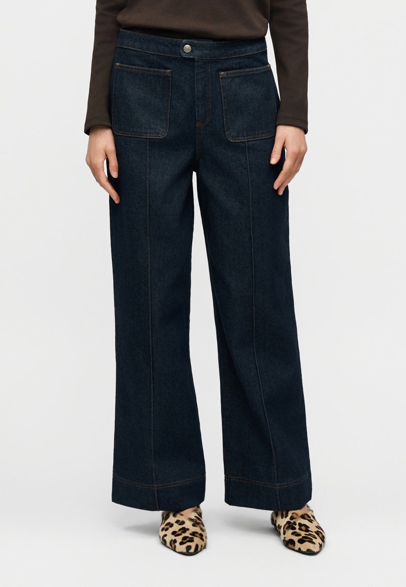 Τζιν σε σκούρο ντένιμ με φαρδιά μπατζάκια, δύο μπροστινές τσέπες, αντίθετη πορτοκαλί ραφή και ψηλή μέση. Το σύνολο ολοκληρώνουν υποδήματα με λεοπάρ μοτίβο.