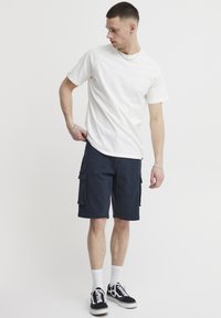 Herreoutfit med en ensfarget hvit bomulls t-skjorte og marineblå cargoshortser med lapper. Tilbehøret inkluderer svarte sneakers med hvite såler.