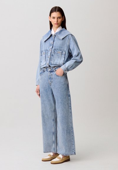 Giacca di denim azzurro chiaro con ampio colletto e tasche frontali, abbinata a jeans azzurri a gamba larga e mocassini beige.