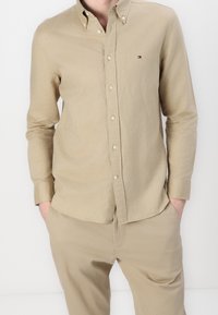 Man in een beige overhemd met lange mouwen en bijpassende beige broek met handen in de zakken tegen een eenvoudige witte achtergrond.