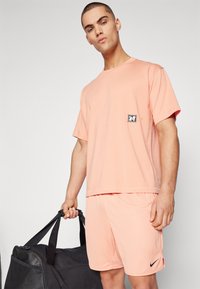 Set sportiv de culoare roz deschis, format dintr-un tricou cu mânecă scurtă și un patch cu logo, împreună cu pantaloni scurți asortați. Modelul ține o geantă de sport neagră.