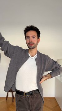 Jonge man binnen in een grijze cardigan over een wit shirt, met een paarse broek en een zwarte riem, poseert met één arm omhoog en de andere op zijn heup.