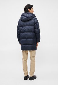 Sötétkék steppelt dzseki kapucnival, steppelt mintázattal és sima felülettel, bézs cargo nadrágra és fekete cipőre viselve.