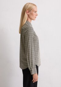 Blouse à motifs noir et blanc avec un col, des manches longues et une coupe ample. Le tissu semble lisse avec un léger éclat.