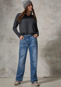 Grauer langärmeliger V-Ausschnitt-Pullover, blaue gerade Jeans, hellgraue Strickmütze. Das Model trägt silberne Sneakers und hat die Hände in den Taschen.