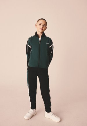 Garçon debout, les mains dans le dos, portant une veste zippée vert foncé et noire, un pantalon noir avec des bandes blanches et des baskets blanches.