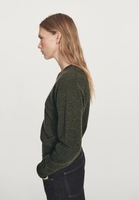Mørkegrøn strikket sweater med rund halsudskæring og lange ærmer, kombineret med mørke denimjeans, set fra siden.