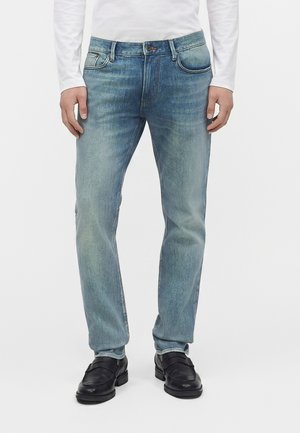 Personne portant un jean bleu délavé, des chaussures en cuir noires et une chemise blanche à manches longues, debout devant un fond blanc uni.