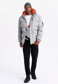 Veste en doudoune grise avec doublure orange, dotée d'une capuche, de poches avant et d'une fermeture éclair. Associée à un t-shirt graphique blanc et à un pantalon cargo noir.
