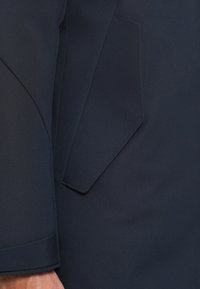 Dunkelblauer Anzugärmel und Klappentaschendetail aus passendem Blazerstoff, mit einer sichtbaren Hand in der unteren linken Ecke.