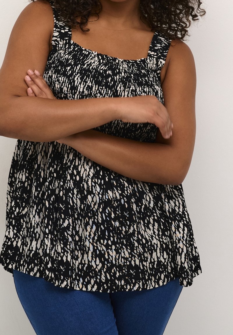 Femme aux cheveux bouclés portant un débardeur noir et blanc à motifs et un jean bleu, bras croisés devant un fond uni.