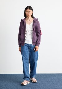 Giovane con lunghi capelli neri, indossando una felpa viola con zip, una maglietta bianca, jeans blu e sneakers beige, in piedi su un tappeto blu.