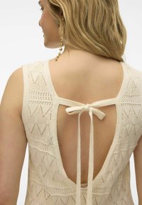 Brede gebreide jurk in crèmekleur met een gestructureerd patroon, diepe V-rug en een gestrikte sluiting bij de nek. Heeft brede bandjes en ingewikkelde details.