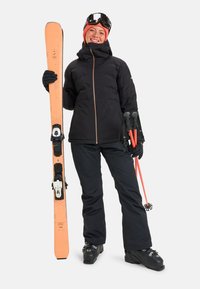 Zwarte pufferjack met ritssluiting, zwarte skibroek, zwarte skischaatsen, houdt oranje skies en oranje stokken vast, skibril op het voorhoofd, glimlachend.