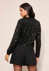 Schwarzer Paillettenpullover mit eingenähtem Strukturstrickdesign, kombiniert mit hochgeschnittenen schwarzen Shorts. Lockere Passform und lange Ärmel, die den Glanz betonen.