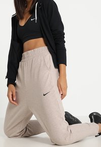 Pantalones de chándal gris claro con una cintura elástica y bolsillos laterales, con un logo de Nike en negro. Combinados con una sudadera con cremallera negra y un top corto.