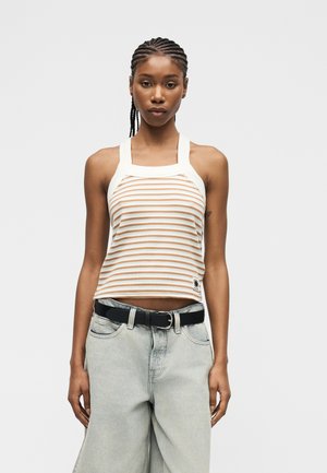 Femeie cu părul împletit, purtând un top tip halter cu dungi albe și bej și blugi largi, de culoare gri deschis, cu o centură neagră, stând în fața unui fundal simplu.