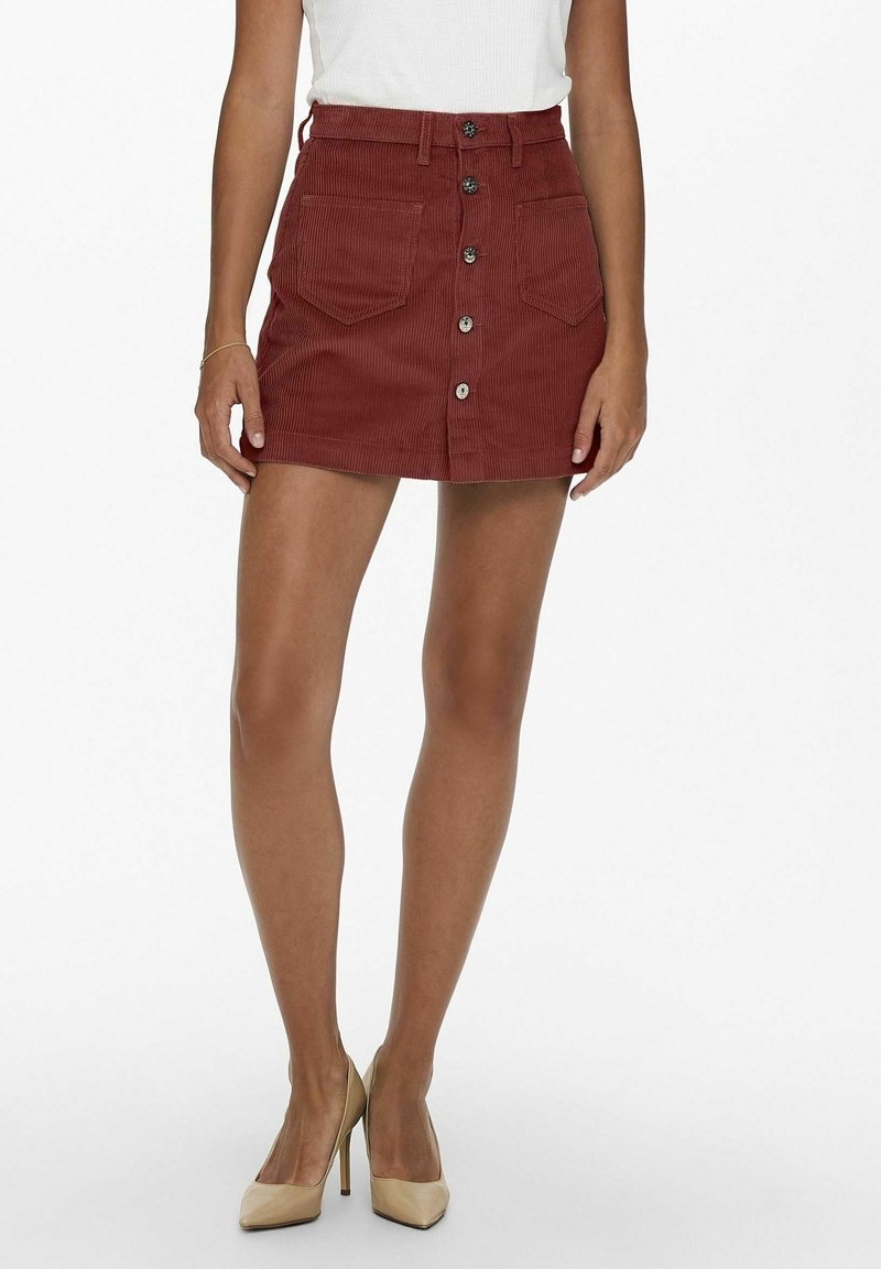 Femme portant une mini-jupe en velours côtelé couleur rouille avec des boutons sur le devant, des poches plaquées, un top blanc sans manches et des talons hauts beiges.