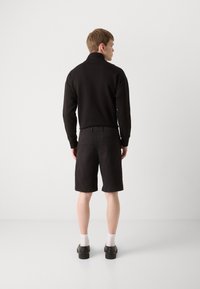Pull noir avec un col montant associé à un short noir, présentant un tissu texturé et des poches arrière. Porté avec des chaussettes blanches et des chaussures noires.