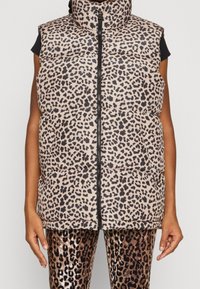 Leopardenmuster-Weste mit Polsterung, hohem Kragen, Reißverschluss und ärmellosem Design. Getragen über passenden Leggings mit Leopardenmuster.