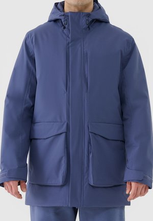 Mann trägt eine lange blaue isolierte Winterjacke mit Kapuze, großen Fronttaschen und Klettverschluss an den Handgelenken, steht vor einem einfarbigen Hintergrund.