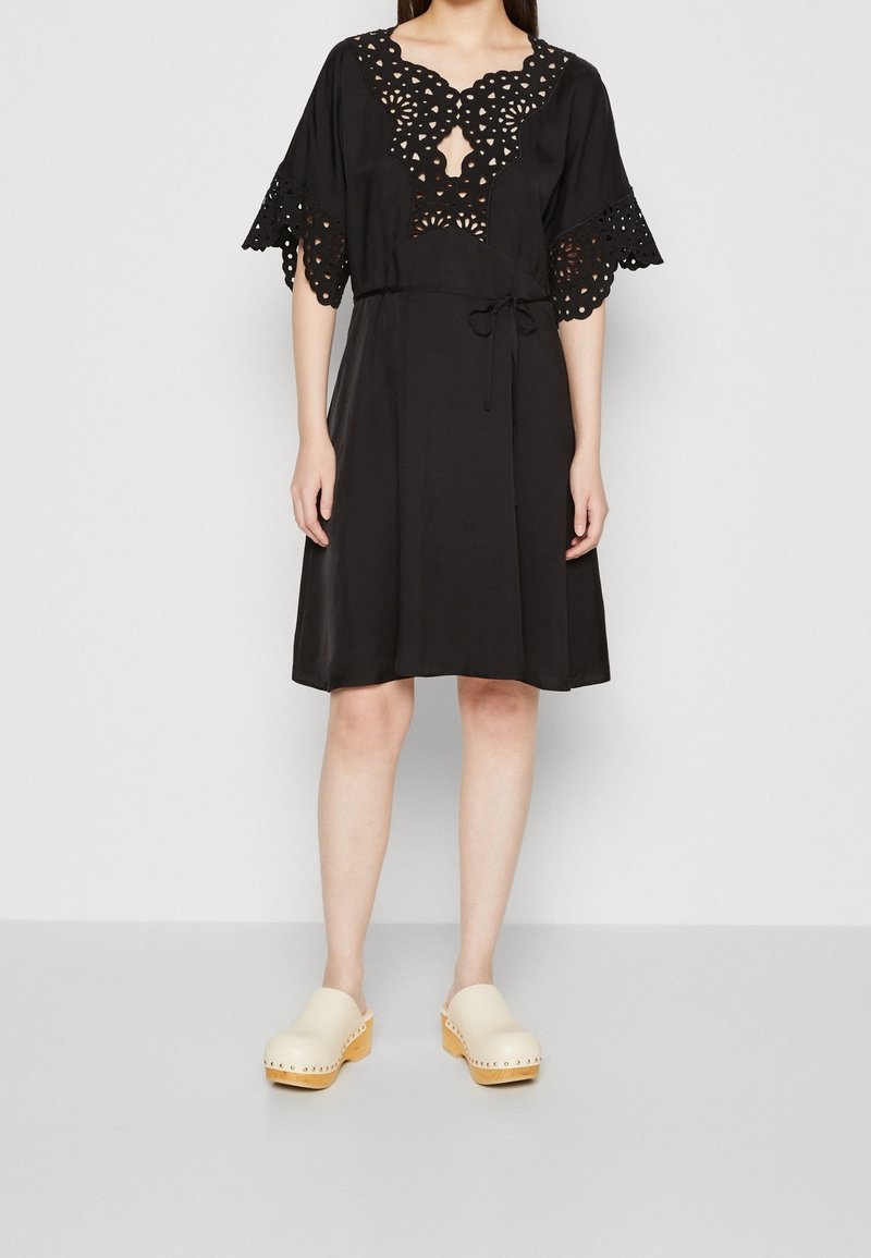 Frau in einem knielangen schwarzen Kleid mit laser-cut Blütenmustern am Ausschnitt und an den Ärmeln, kombiniert mit beigen Holzclogs.