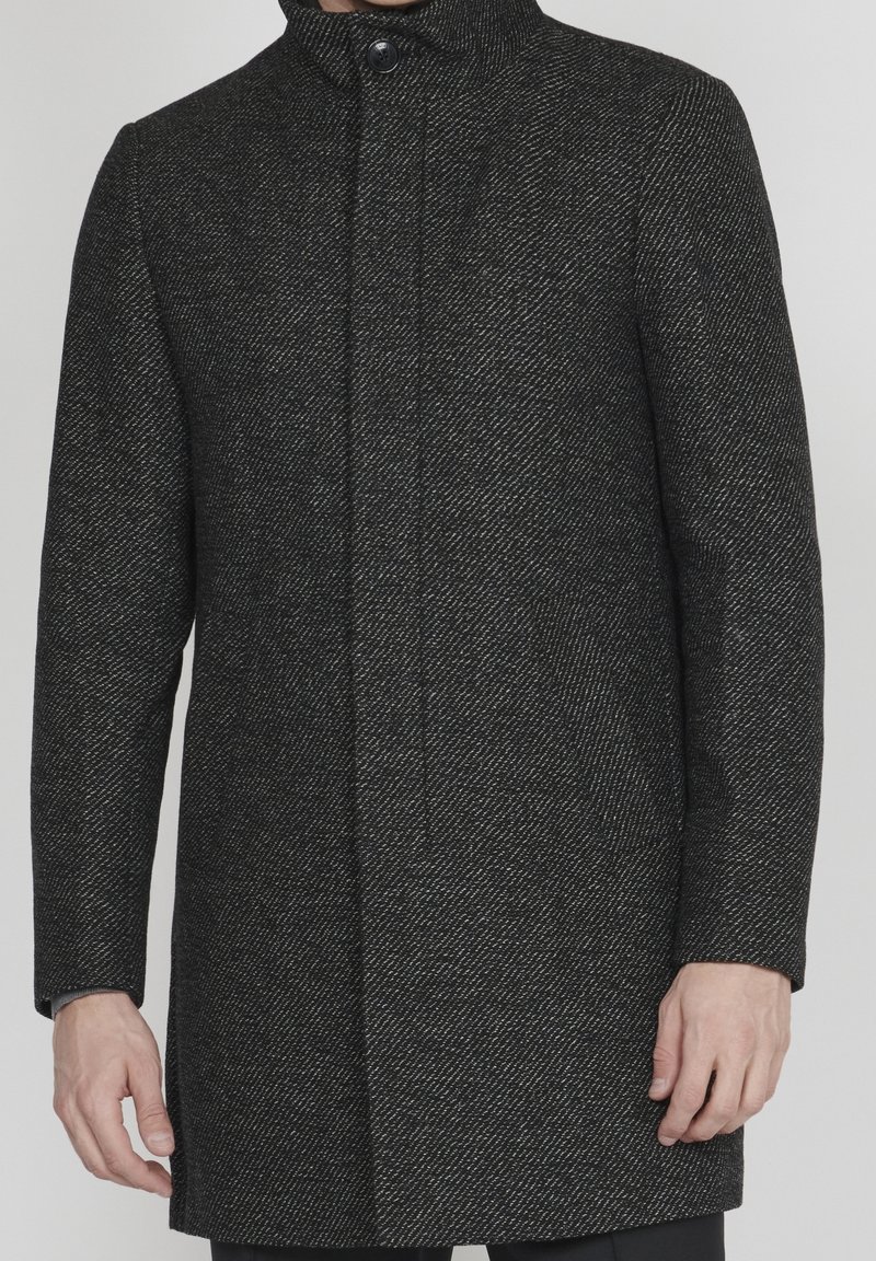 Czarny i szary płaszcz w jodełkę z wysokim kołnierzem, zapięciem na jeden guzik i bocznymi kieszeniami; tkana tekstura nadaje głębi projektowi.