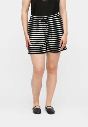 Vrouw die zwart-wit gestreepte shorts draagt met een trekkoord in de taille, een zwart topje en zwarte Mary Jane platte schoenen, staand tegen een effen achtergrond.