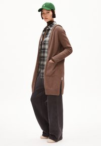 Brauner langer Cardigan mit Taschen über einem karrierten Hemd, kombiniert mit lockeren, dunklen Hosen und einer grünen Mütze. Weiche Stricktextur, lässige Passform.