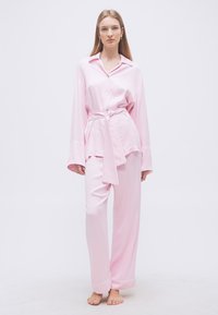Γυναίκα που στέκεται ξυπόλητη φορώντας χαλαρή σατέν πιτζάμα ανοιχτόροζ, με πάνω μέρος με κουμπιά και μακριά μανίκια, δεμένο στη μέση, και παντελόνι με φαρδιά μπατζάκια.