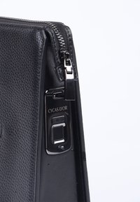 Sort lædertaske med en tekstureret overflade, der har en sølv lynlås og et enhedsdisplay med ind-/udgangsport og brandnavn.