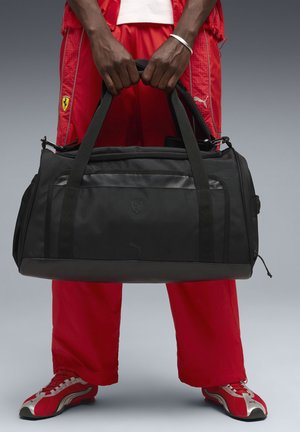 Svart duffelväska med robust tyg, dubbla handtag och en framlogotyp. Märkbara texturerade detaljer och en solid botten.