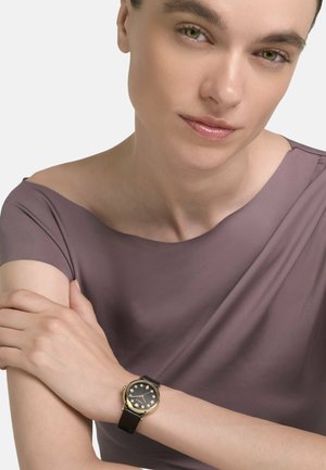 Mujer con cabello corto que lleva una blusa malva con los hombros descubiertos y un reloj de pulsera de cuero negro con caja dorada y marcadores de hora de cristal.