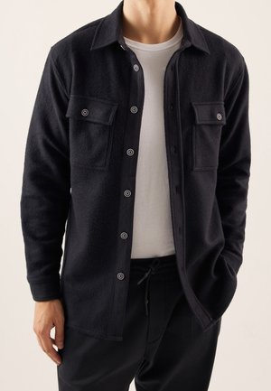 Homme portant une veste-chemise noire boutonnée avec deux poches poitrine sur un T-shirt blanc et un pantalon noir, une main dans la poche.