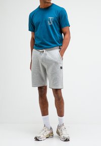 Teal katoenen t-shirt met een grafisch ontwerp op de zak, gecombineerd met grijze shorts. De shorts hebben een elastische tailleband en zijzakken.