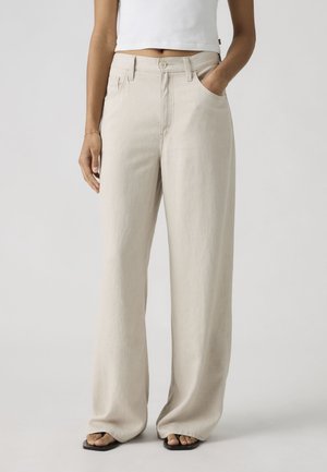 Oseba v ohlapnih bež hlačah in črnih sandalih, z eno roko v žepu in krajšim belim vrhom, stoji ob enobarvnem ozadju.