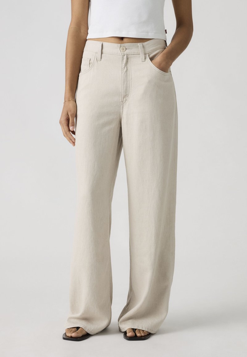 Person iført løse beige bukser og sorte sandaler, med den ene hånd i lommen og en kort hvid top, stående mod en ensfarvet baggrund.