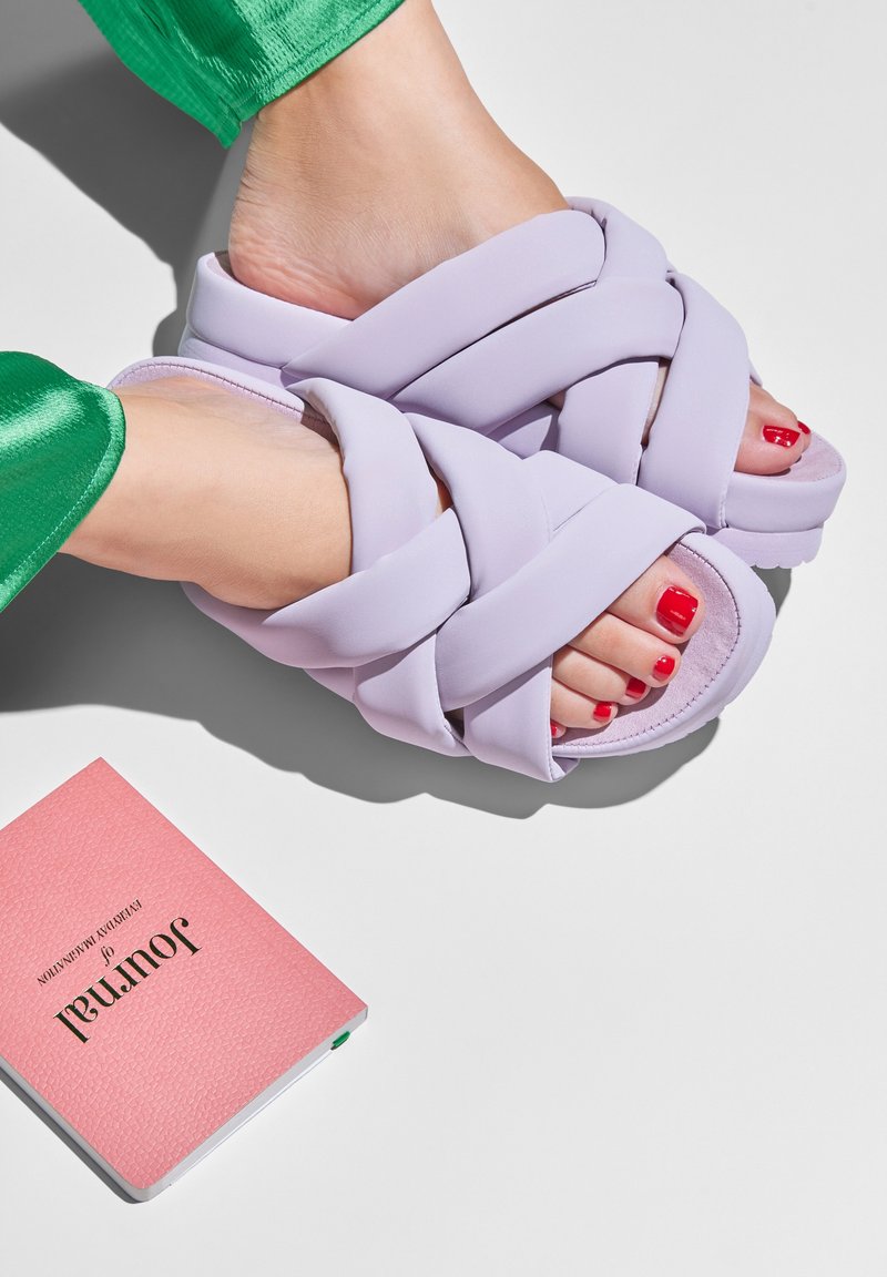 Lavender cross-band slide sandals made from soft material. Notable details include a textured upper and red toenail polish. A pink notebook is nearby.