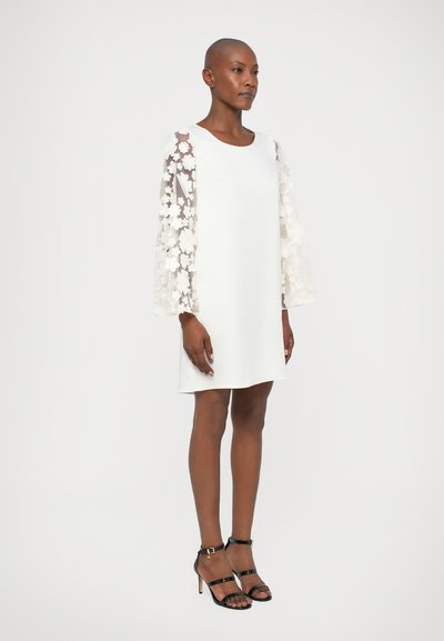 Vestido blanco con mangas transparentes de estampado floral; forma en A, textura suave. Llevado con tacones negros con tiras.