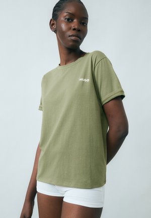 Femme portant un t-shirt HUGO vert olive et un short blanc, debout avec une expression neutre devant un fond clair.