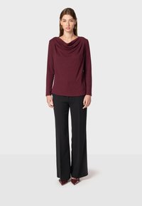 Bordowy top z długim rękawem i drapowanym dekoltem, połączony z czarnymi spodniami o szerokich nogawkach. Modelka uzupełniła stylizację złotymi kolczykami i bordowymi butami.