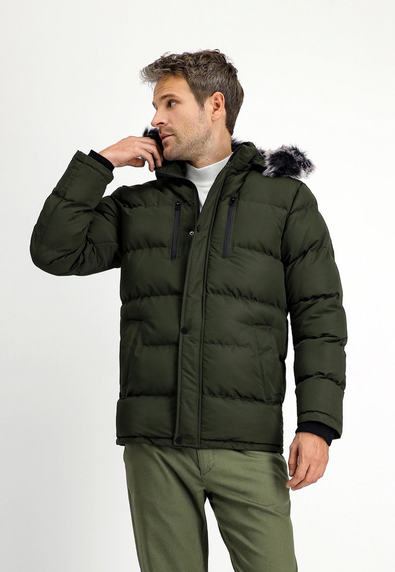 Chaqueta tipo puffer de color verde oscuro con capucha, que presenta un ribete de piel sintética, bolsillos con cremallera y una textura acolchada. Llevada con una camisa blanca y pantalones verdes.