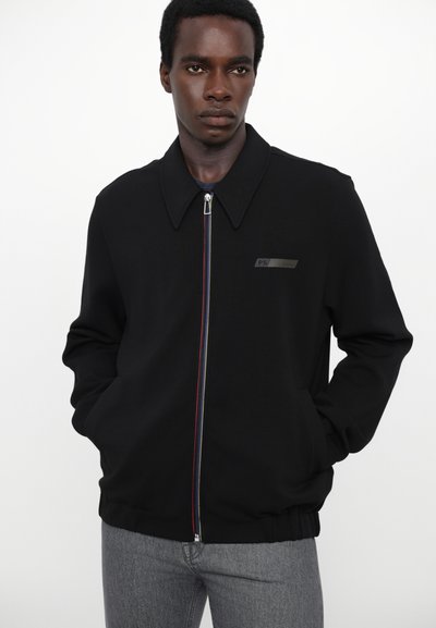 Veste noire zippée avec un large col, arborant un logo subtil et des accents de fermeture éclair multicolores. Portée sur un jean gris ajusté.
