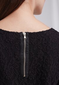 Svart spetsklänning med ett blommönster, med en synlig silverfärgad dragkedja på baksidan och strukturdetaljer i tyget.