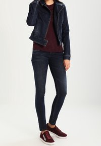 Dark denim jacket, maroon textured top, dark skinny jeans, and burgundy sneakers with white accents. Casual outfit on a plain background.