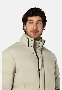 Lichtbeige pufferjas met hoge kraag, zwarte rits en drukknopen. Gladde textuur, met horizontale doorgestikte vulling.