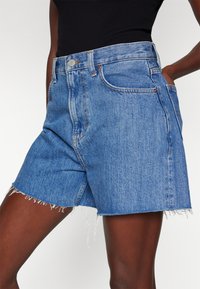 Bermudas de mezclilla en un lavado azul claro con cintura alta, dobladillo deshilachado y diseño clásico de cinco bolsillos. Presenta acentos de hardware en tono cobre.